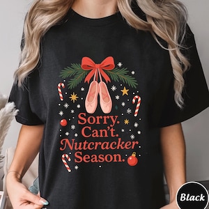 Op de afbeelding: Zwart T-shirt met een feestelijk ontwerp. De afbeelding bevat een rode strik, balletschoenen, zuurstokken, sterren en de tekst "Sorry. Can't. Nutcracker. Season."