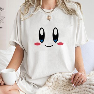 May include: Cream-colored t-shirt featuring a cartoon face with large black eyes, blue pupils, and pink cheeks. The design is simple and playful, perfect for casual wear. The shirt has short sleeves and a relaxed fit.