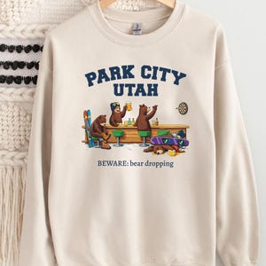 Può includere: Felpa beige con grafica di orsi in un bar, la scritta "PARK CITY UTAH" in blu navy e la frase "BEWARE: bear dropping" in nero. Gli orsi bevono birra e uno è svenuto.
