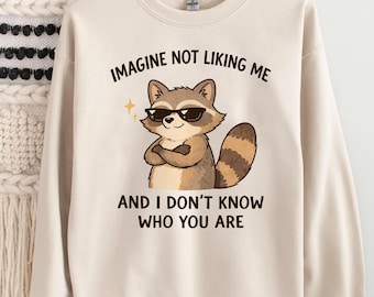 Può includere: Felpa color crema con la scritta "IMAGINE NOT LIKING ME AND I DON'T KNOW WHO YOU ARE". La grafica presenta un procione con occhiali da sole e le braccia incrociate. Il procione è marrone, beige e bianco.