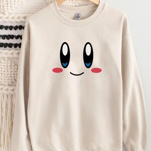 May include: A cream-colored sweatshirt featuring a cartoon face with large black and white eyes, blue pupils, a small smile, and pink cheeks. The design is centered on the front of the long-sleeved crew neck top.