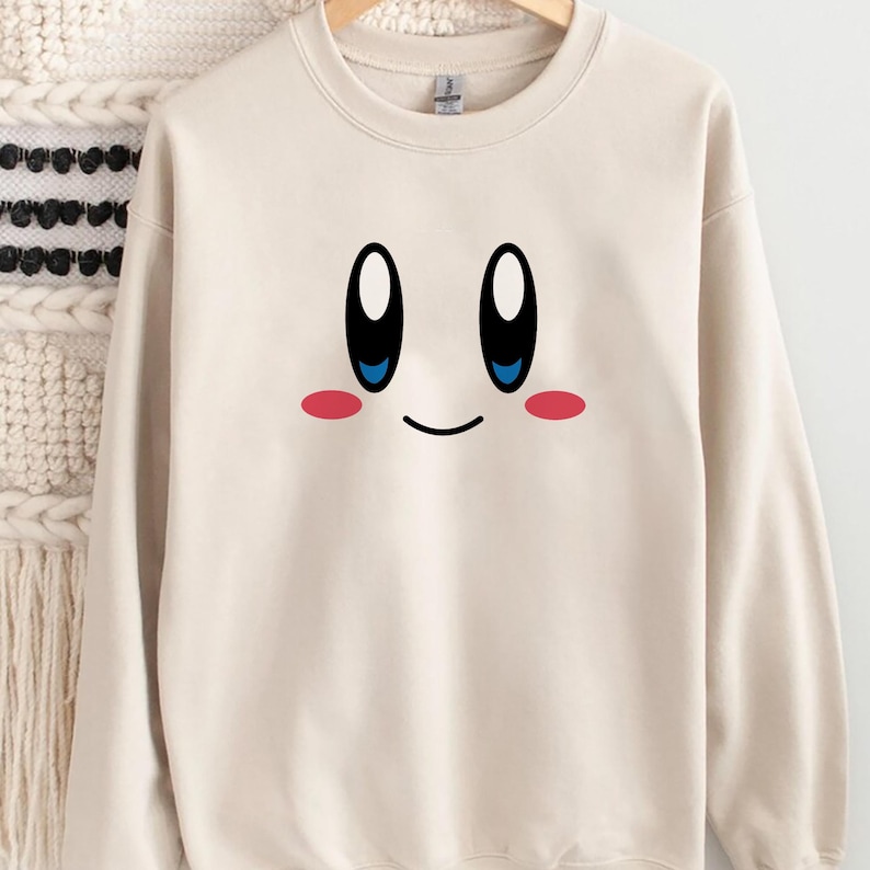 May include: A cream-colored sweatshirt featuring a cartoon face with large black and white eyes, blue pupils, a small smile, and pink cheeks. The design is centered on the front of the long-sleeved crew neck top.
