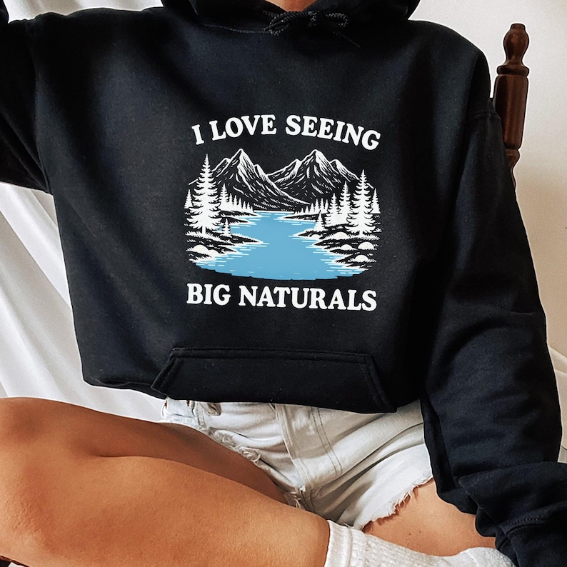 May include: Black hoodie with a front pocket and a drawstring hood. The graphic on the front features a mountain and lake scene with the text "I LOVE SEEING BIG NATURALS" in white.