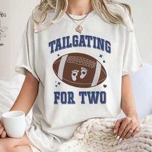 Peut inclure: T-shirt blanc avec un graphique de ballon de football bleu et marron et le texte "TAILGATING FOR TWO".
