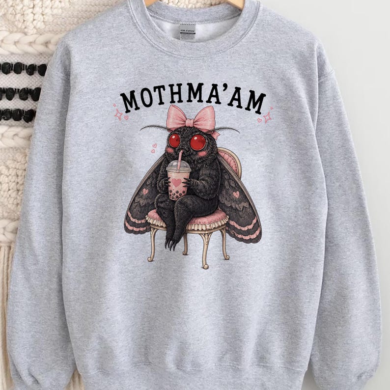 May include: A light gray crewneck sweatshirt featuring a graphic of a black moth with red eyes, wearing a pink bow, sitting on a small stool, and holding a bubble tea. The text MOTHMA'AM is printed in black above the moth illustration.