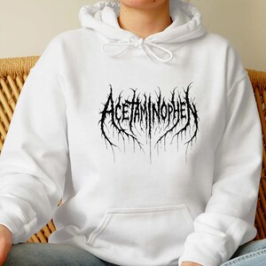 Puede incluir: Sudadera con capucha blanca con la palabra "Acetaminophen" en negro, en una fuente g&oacute;tica. El texto tiene un efecto desgastado y texturizado. La sudadera tiene una capucha con cord&oacute;n y un bolsillo delantero.