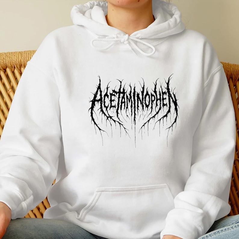Puede incluir: Sudadera con capucha blanca con la palabra "Acetaminophen" en negro, en una fuente g&oacute;tica. El texto tiene un efecto desgastado y texturizado. La sudadera tiene una capucha con cord&oacute;n y un bolsillo delantero.