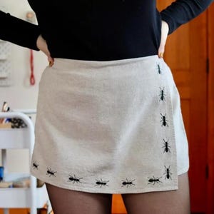 May include: A beige mini skirt featuring a wrap-style front panel with a series of small, black embroidered ant motifs arranged along the bottom hem and up the vertical edge of the overlapping fabric section.