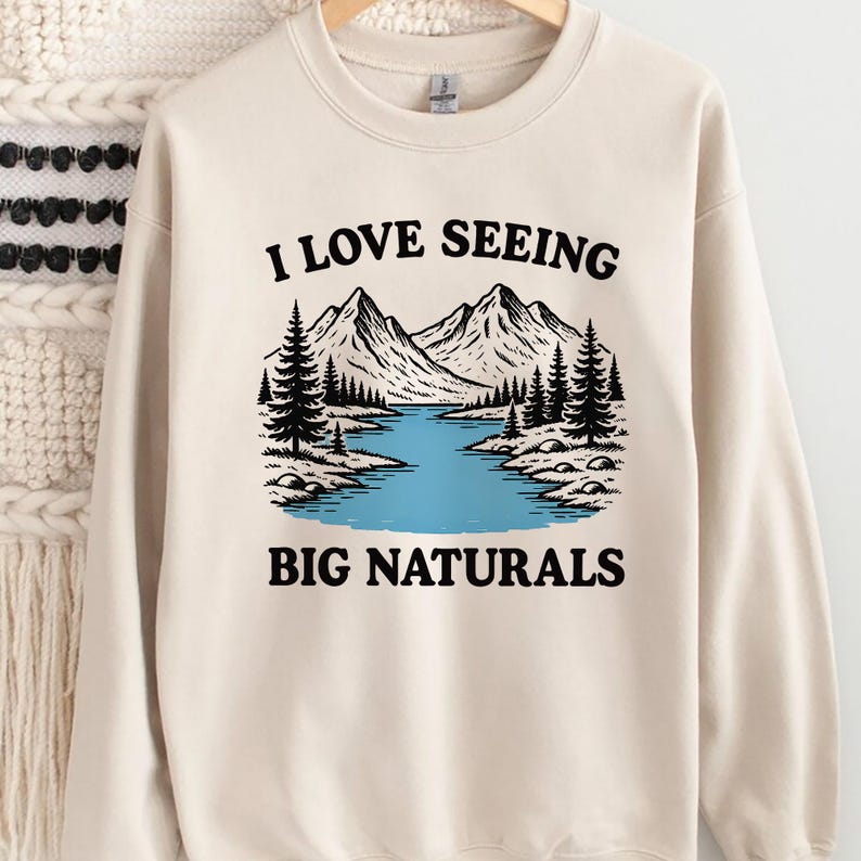 May include: Cream-colored sweatshirt with a graphic of a mountain range, river, and trees. The text "I LOVE SEEING BIG NATURALS" is printed above and below the graphic in black.