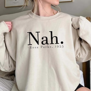 Peut inclure: Un sweat-shirt beige avec le texte "Nah." et "Rosa Parks, 1955" imprimé sur le devant.