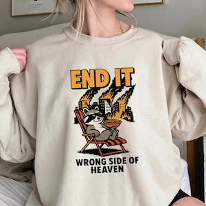 Puede incluir: Sudadera beige con un gráfico que presenta un mapache con gafas de sol relajándose en una silla de playa, con edificios en llamas al fondo. El texto "END IT" está encima del mapache y "WRONG SIDE OF HEAVEN" debajo.
