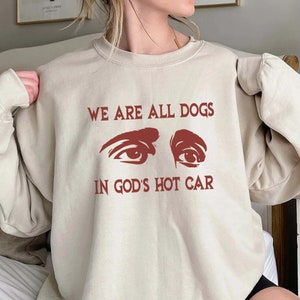 Può includere: Una felpa beige con la scritta "WE ARE ALL DOGS IN GOD'S HOT CAR" stampata in rosso. Il testo è accompagnato da una grafica di due occhi.