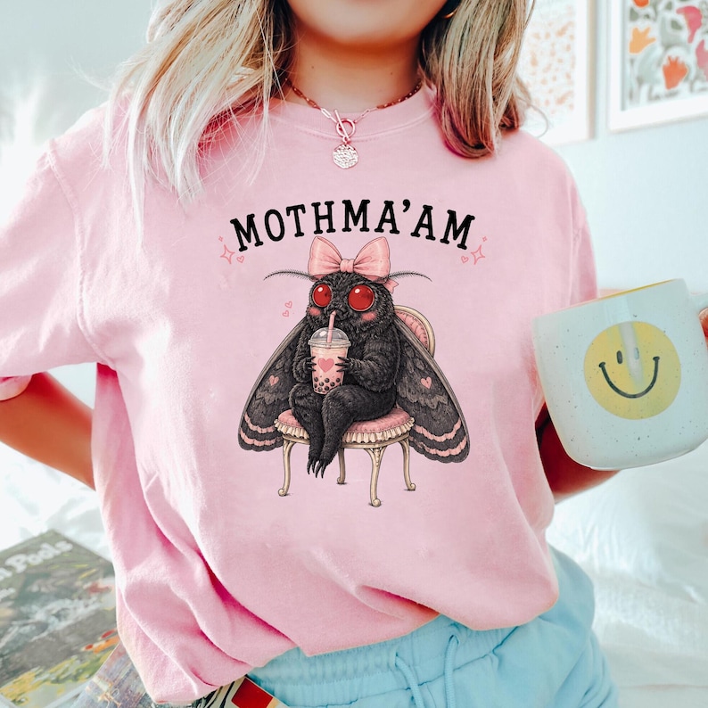 May include: A light pink t-shirt featuring a graphic of a black moth with red eyes, wearing a pink bow, sitting on a stool, and holding a boba tea drink. The text MOTHMA'AM is printed in black above the moth, accompanied by small pink heart icons.