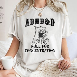 May include: White t-shirt with a black and white illustration of a cat biting a twenty-sided die. The text "ADHD&D" is above the cat and "ROLL FOR CONCENTRATION" is below the cat.