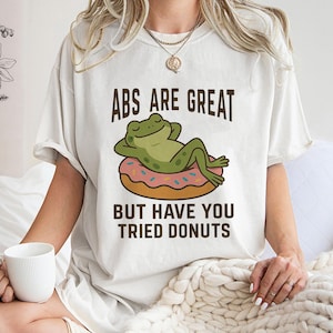 May include: Cream-coloured t-shirt with the text "ABS ARE GREAT BUT HAVE YOU TRIED DONUTS". The graphic features a green frog relaxing on a pink frosted donut with brown sprinkles. The shirt has short sleeves.