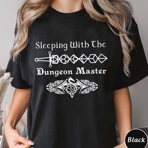 May include: Black t-shirt with white text that reads "Sleeping With The Dungeon Master" and a graphic of a sword, dice, and a dragon.