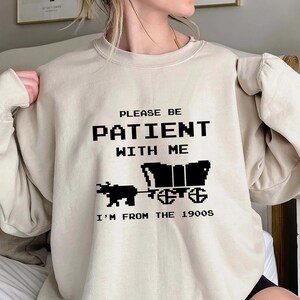 May include: A beige sweatshirt with a black graphic print that says "Please be patient with me I'm from the 1900s". The graphic features a pixelated image of a wagon pulled by an ox.