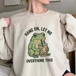 May include: Beige sweatshirt featuring a green frog drinking bubble tea. The text "HANG ON, LET ME OVERTHINK THIS" is printed above and below the frog in black. The frog graphic is holding a clear cup with brown boba.