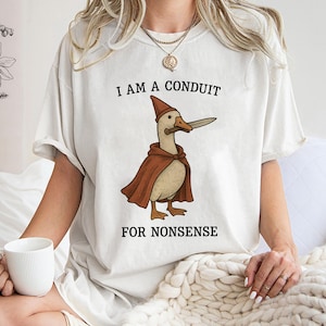 May include: Cream-colored t-shirt featuring a cartoon goose in a red cape and hat, holding a knife. The text "I AM A CONDUIT FOR NONSENSE" is printed above and below the goose illustration. The shirt has short sleeves.