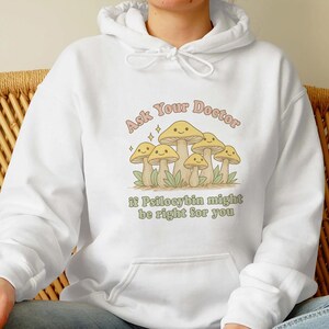 K&ouml;nnte beinhalten: Wei&szlig;er Kapuzenpullover mit einem Pilz-Motiv und dem Text "Ask Your Doctor if Psilocybin might be right for you". Das Design zeigt Cartoon-Pilze mit l&auml;chelnden Gesichtern.