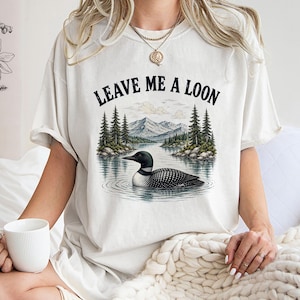 May include: An off-white short-sleeve t-shirt featuring a graphic print of a loon swimming on a lake with pine trees and mountains in the background. The text "LEAVE ME A LOON" is printed in black capital letters above the nature illustration.