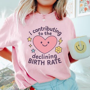 May include: A light pink t-shirt with the text "I contributing to the declining BIRTH RATE" and a smiling pink heart graphic. The shirt is paired with a white mug featuring a yellow smiley face.