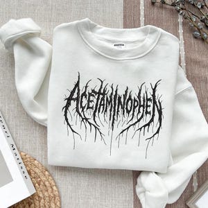 May include: White crewneck sweatshirt with the word "Acetaminophen" in a black, gothic-style font. The text has a distressed, textured appearance, with jagged lines extending from the letters.