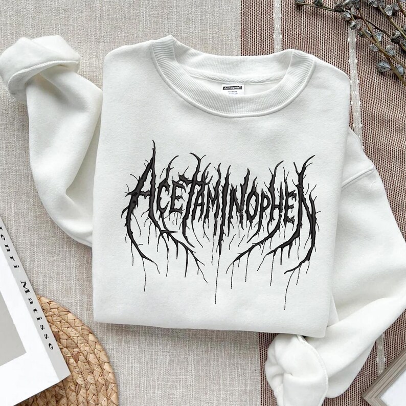 May include: White crewneck sweatshirt with the word "Acetaminophen" in a black, gothic-style font. The text has a distressed, textured appearance, with jagged lines extending from the letters.