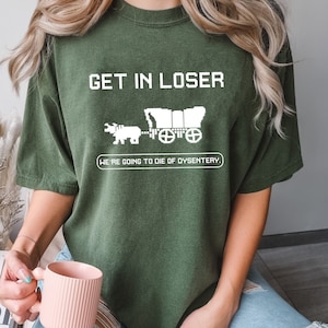 May include: A green t-shirt with the text "GET IN LOSER, WE'RE GOING TO DIE OF DYSENTERY." in white. The text is in a pixelated font. A pixelated image of a covered wagon pulled by an ox is above the text.