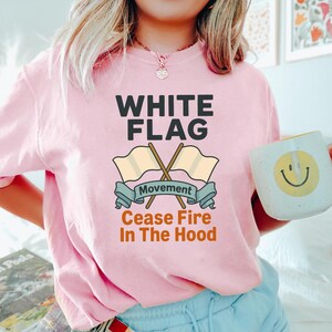 May include: A light pink t-shirt with the words "WHITE FLAG" in bold black letters. Below the text are two crossed beige flags with the word "Movement" on a blue banner. The phrase "Cease Fire In The Hood" is printed below.