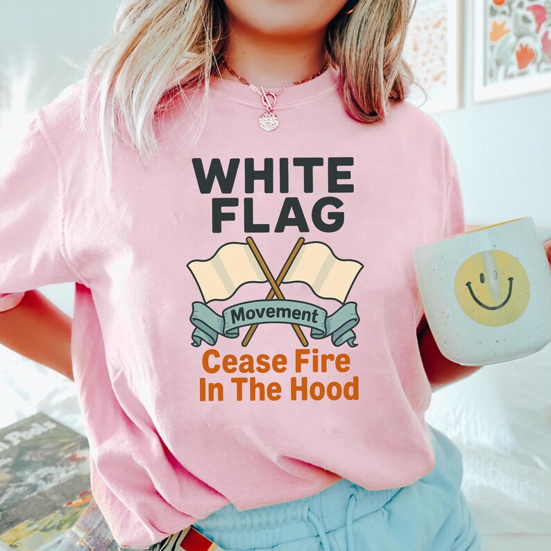 May include: A light pink t-shirt with the words "WHITE FLAG" in bold black letters. Below the text are two crossed beige flags with the word "Movement" on a blue banner. The phrase "Cease Fire In The Hood" is printed below.