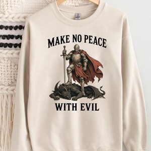 May include: Cream-colored sweatshirt featuring a graphic of a knight standing on a dragon, holding a sword. The text "MAKE NO PEACE WITH EVIL" is printed above and below the illustration in black.