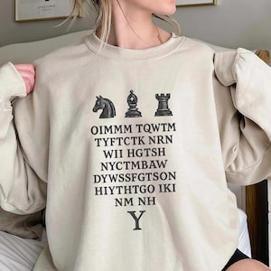 Könnte beinhalten: Beiges Sweatshirt mit schwarzen Schachfiguren: ein Springer, ein Läufer und ein Turm. Darunter befinden sich mehrere Textzeilen, die mit dem Buchstaben "Y" enden. Das Sweatshirt hat eine neutrale Farbe.