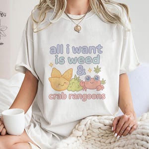 Op de afbeelding: Crèmekleurig T-shirt met de tekst "all I want is weed & crab rangoon" in pastelkleuren. De afbeelding bevat een lachende krab, een cannabisblad en een wonton.