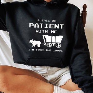 May include: Black hooded sweatshirt with white text that reads "Please be patient with me I'm from the 1900s". The text is in a pixelated font and there is a pixelated image of a covered wagon pulled by an ox.