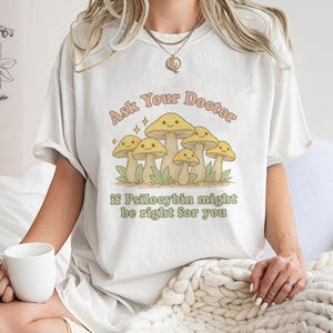 K&ouml;nnte beinhalten: Cremefarbenes T-Shirt mit einer Grafik von l&auml;chelnden Pilzen und dem Text "Ask Your Doctor if Psilocybin might be right for you". Das Design verwendet Gelb- und Hellgr&uuml;nt&ouml;ne und eine Retro-Schriftart.
