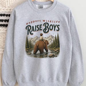 May include: A pale grey sweatshirt featuring the text "Support Wildlife Raise Boys" above an illustration of a boy riding a brown bear in a river. The background shows mountains and trees. The boy wears light-coloured clothing.