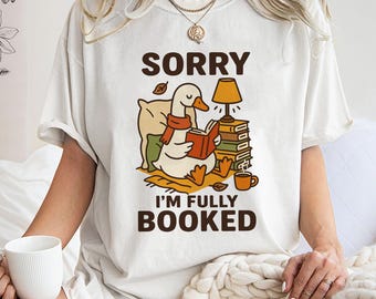 May include: A cream-coloured t-shirt with the text "SORRY I'M FULLY BOOKED." The graphic depicts a cartoon goose reading a book, wearing a scarf, and sitting near a stack of books and a lamp. A coffee cup is also present.