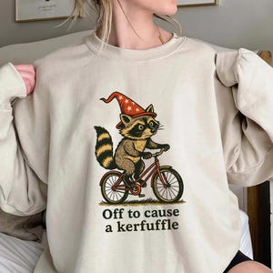 Könnte beinhalten: Beiges Sweatshirt mit einem Cartoon-Waschbären, der einen roten Zaubererhut mit Sternen trägt und ein rotes Fahrrad fährt. Der Text "Off to cause a kerfuffle" ist darunter gedruckt.