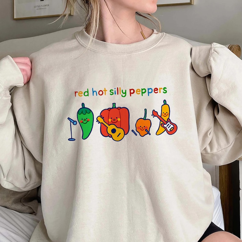 Pu&ograve; includere: Una felpa beige chiaro con una grafica di peperoni dei cartoni animati che suonano musica. Il testo "red hot silly peppers" &egrave; stampato sulla felpa.