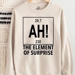 May include: A cream-coloured sweatshirt featuring a black graphic. The graphic is a square with the numbers "29,7" and "230" at the top and bottom, and the letters "AH!" in the centre. Below the square, the text reads "THE ELEMENT OF SURPRISE."