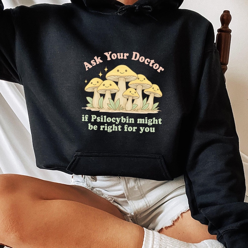K&ouml;nnte beinhalten: Schwarzer Kapuzenpullover mit einer Fronttasche und einer Grafik von l&auml;chelnden Pilzen mit dem Text "Ask Your Doctor if Psilocybin might be right for you" in Rosa und Gr&uuml;n.