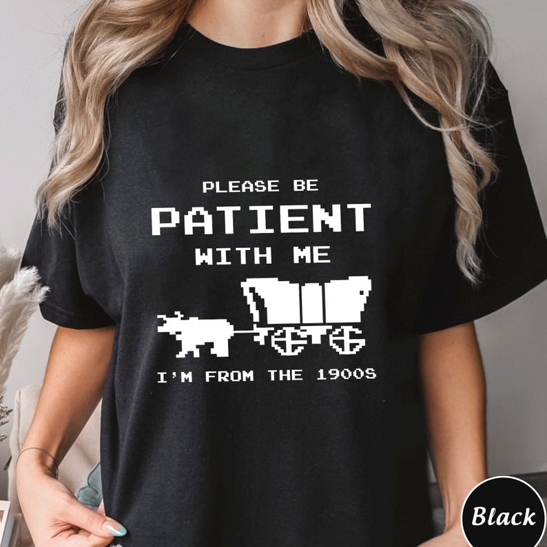 May include: Black t-shirt with white text that reads "Please be patient with me I'm from the 1900s". The text is in a pixelated font and there is a pixelated image of a covered wagon being pulled by an ox.