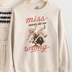 May include: Cream-colored sweatshirt with a graphic of a cowgirl riding a bull, with the words "miss never do no wrong" in a reddish-brown script. The cowgirl is wearing a cowboy hat, boots, and denim shorts.