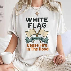 May include: Cream-colored t-shirt with the text "WHITE FLAG" above a graphic of two crossed flags and a banner that reads "Movement" and "Cease Fire In The Hood". The shirt is worn by a person holding a white mug.