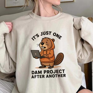 May include: Beige sweatshirt with the text "IT'S JUST ONE DAM PROJECT AFTER ANOTHER." The graphic features a cartoon beaver wearing an orange hoodie and holding a laptop. The beaver has a large tail and is smiling.
