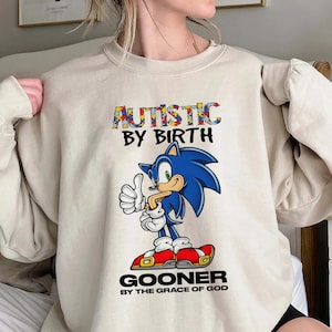 Könnte beinhalten: Beiges Sweatshirt mit dem Text "AUTISTIC BY BIRTH" in bunten Buchstaben und einem Cartoon-Bild von Sonic the Hedgehog, der den Daumen hochhält. Unter dem Bild steht der Text "GOONER BY THE GRACE OF GOD".