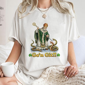 Może przedstawiać: Kremowa koszulka z grafiką św. Patryka z laską, zielonymi szatami i czerwonym kapeluszem, walczącego z wężami. Tekst "Go'n Git!!" jest wydrukowany pod obrazem, z koniczyną.