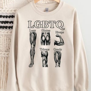 Op de afbeelding: Crèmekleurige sweatshirt met het woord "LGBTQ" in zwarte blokletters boven anatomische illustraties van spieren. Daaronder staan de woorden "Legs, Glutes, Biceps, Triceps, Quads".