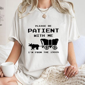 May include: White t-shirt with black text that reads "Please be patient with me I'm from the 1900s". The text is in a pixelated font and there is a pixelated image of a wagon pulled by an ox.